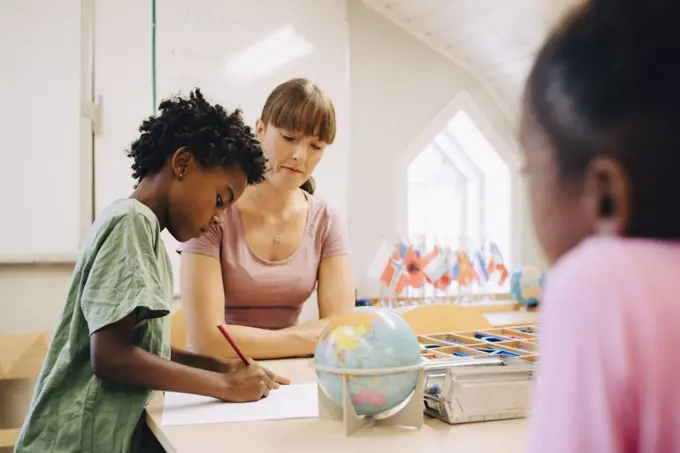 Teacher looking at boy writing on paper at table in classroom