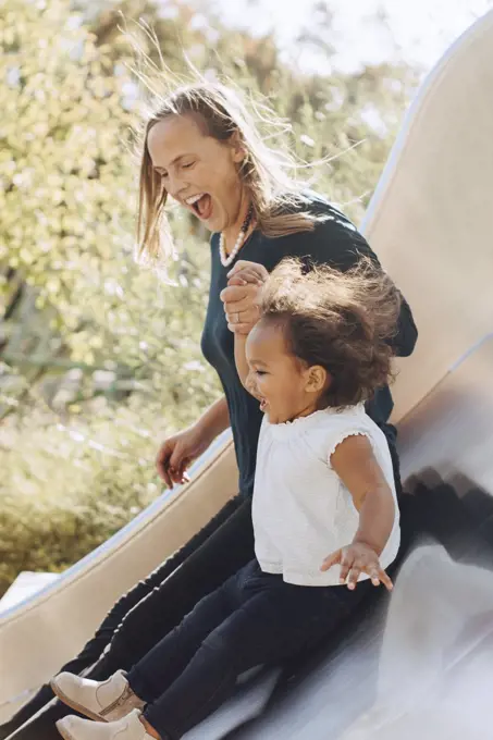 Happy mother sliding with daughter on slide at park