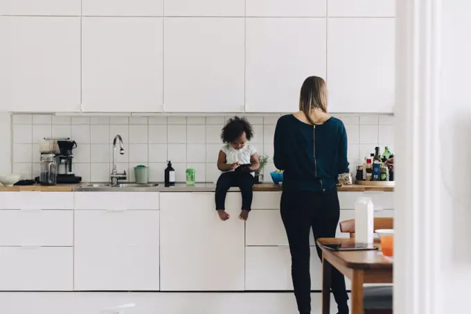 Full length of girl using smart phone while mother working in kitchen at home