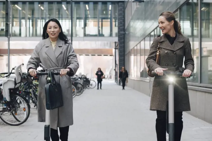 Smiling female colleagues riding electric push scooter on footpath by building in city