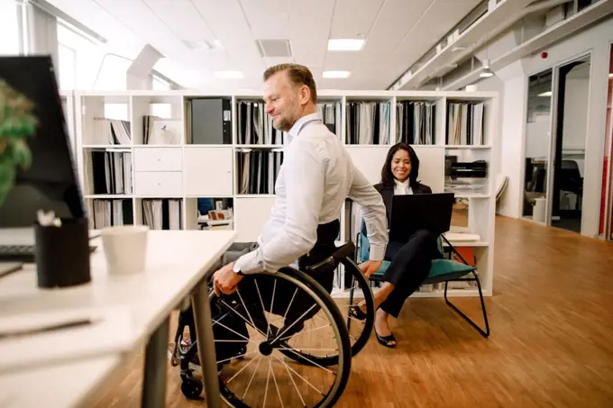 Disabled male professional sitting on wheelchair while smiling businesswoman using laptop at work place