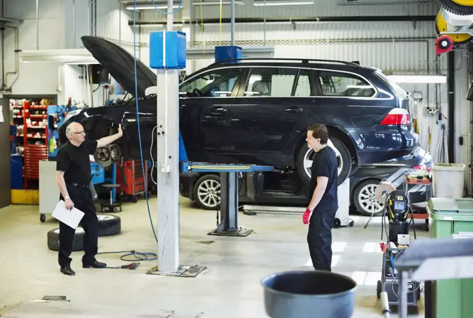 Male mechanics discussing while standing against car jack at workshop