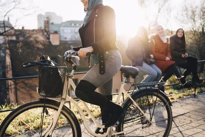 Low section of woman cycling by female friends sitting on railing in city