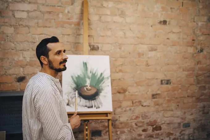 Side view of male artist standing by painting against brick wall at studio