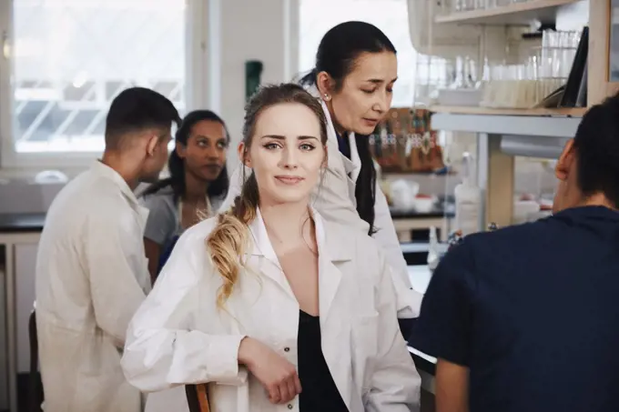 Portrait of confident young female student sitting against teacher friends in chemistry laboratory