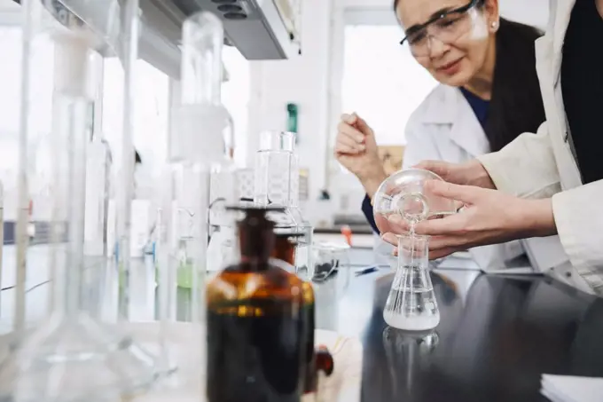 Midsection of young female student pouring solution in flask by teacher at chemistry laboratory