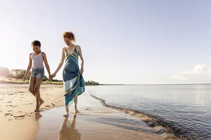 Full length of woman holding hands with daughter while walking on shore at beach against sky