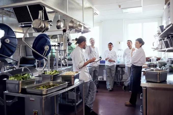 Male chef holding digital tablet while discussing with colleagues in commercial kitchen