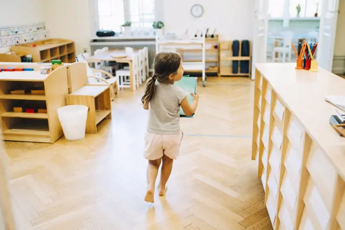 Full length rear view of girl running with book in classroom at kindergarten