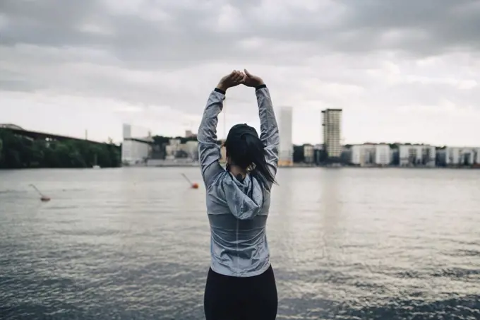 Rear view of female athlete stretching arms while standing against sea in city