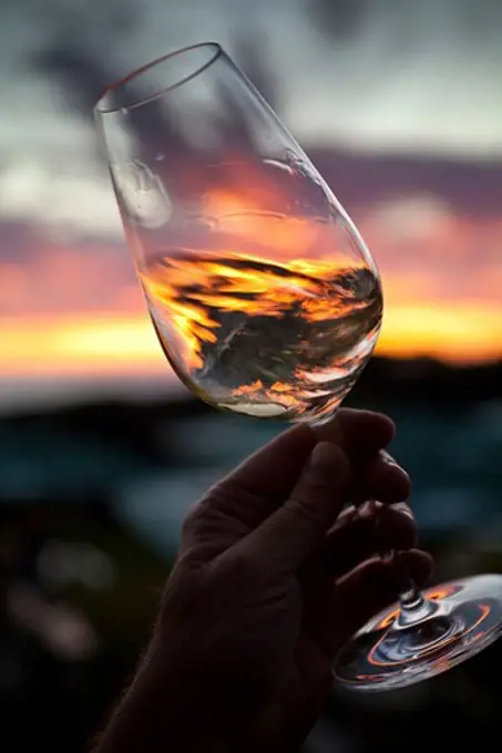 Hand holding and swirling glass of white wine against sunset, Stellenbosch, Western Cape, South Africa