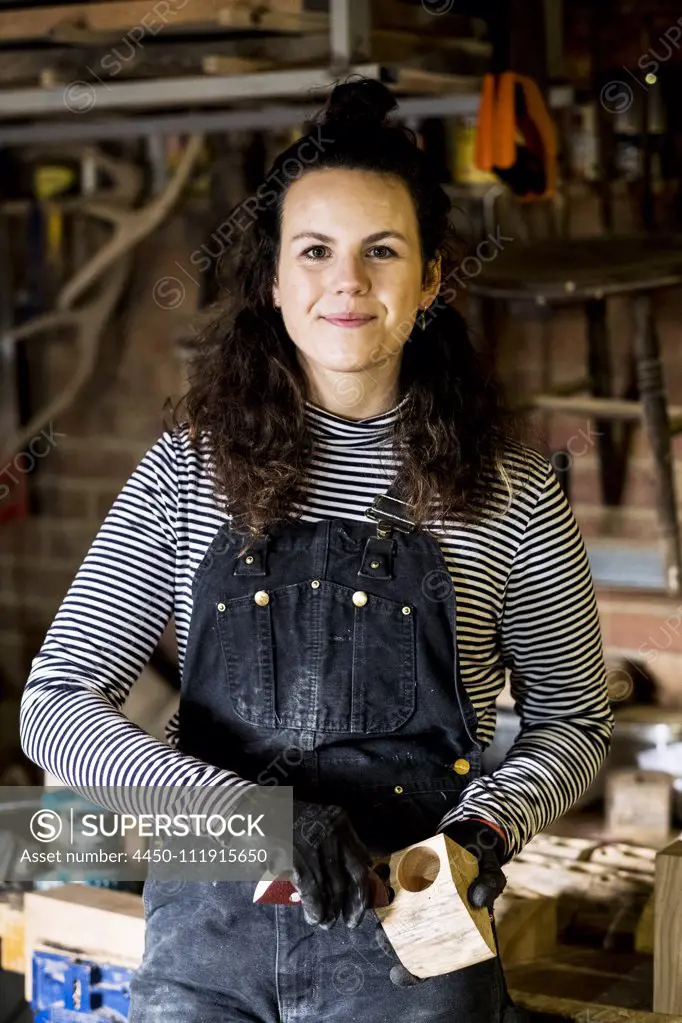 Woman with long brown hair wearing dungarees standing in wood workshop, smiling at camera.