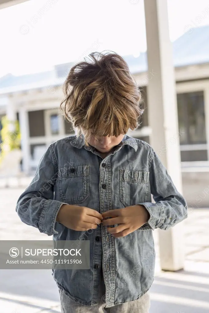 A young boy buttoning up his denim shirt