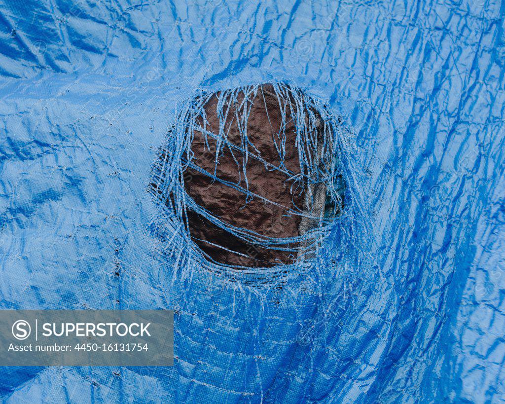 Close up worn blue tarp with tattered hole in center - SuperStock