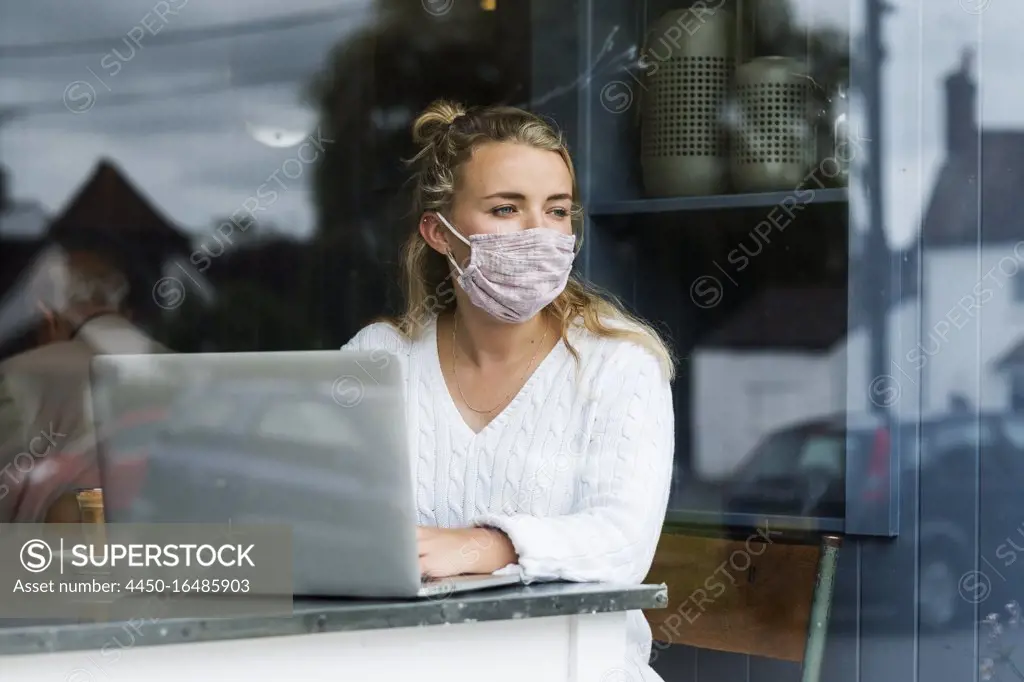 Woman wearing face mask sitting alone at a cafe table with a laptop computer, working remotely.