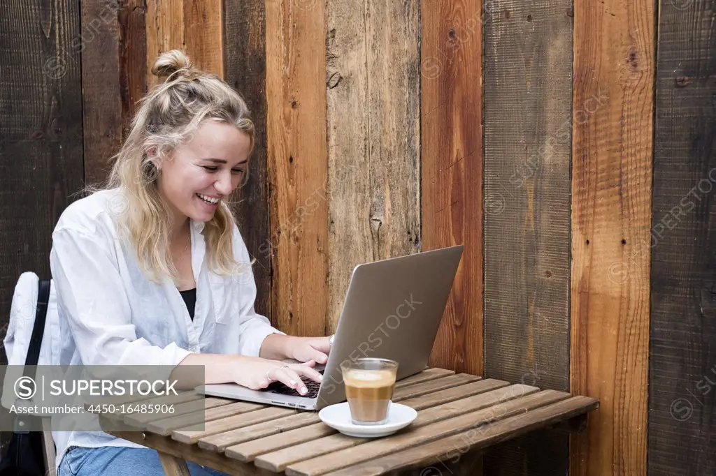 Young blond woman sitting alone at a cafe table with a laptop computer, working remotely.