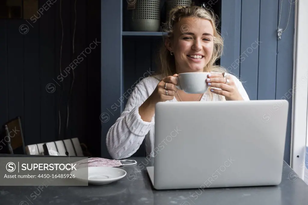 Young blond woman sitting alone at a cafe table with a laptop computer, working remotely.