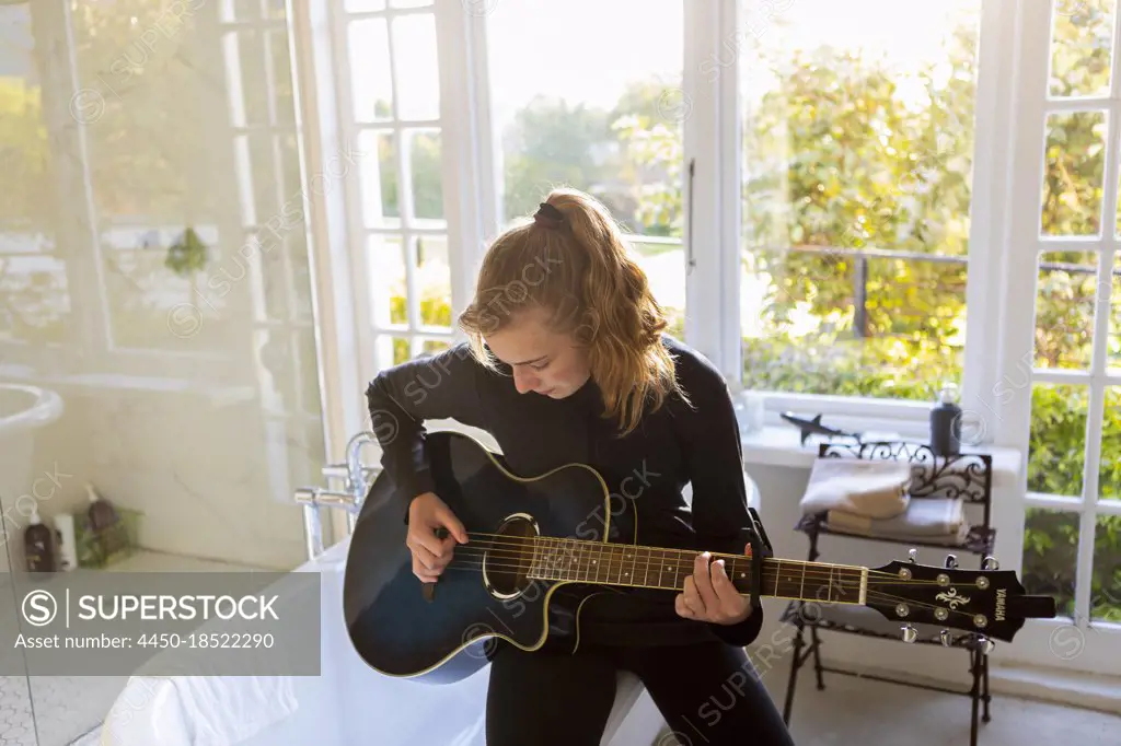 Teenage girl seated on the edge of a bathtub, playing accoustic guitar. 