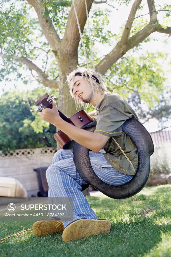 Man playing guitar sitting on a tyre swing in a garden, singing.