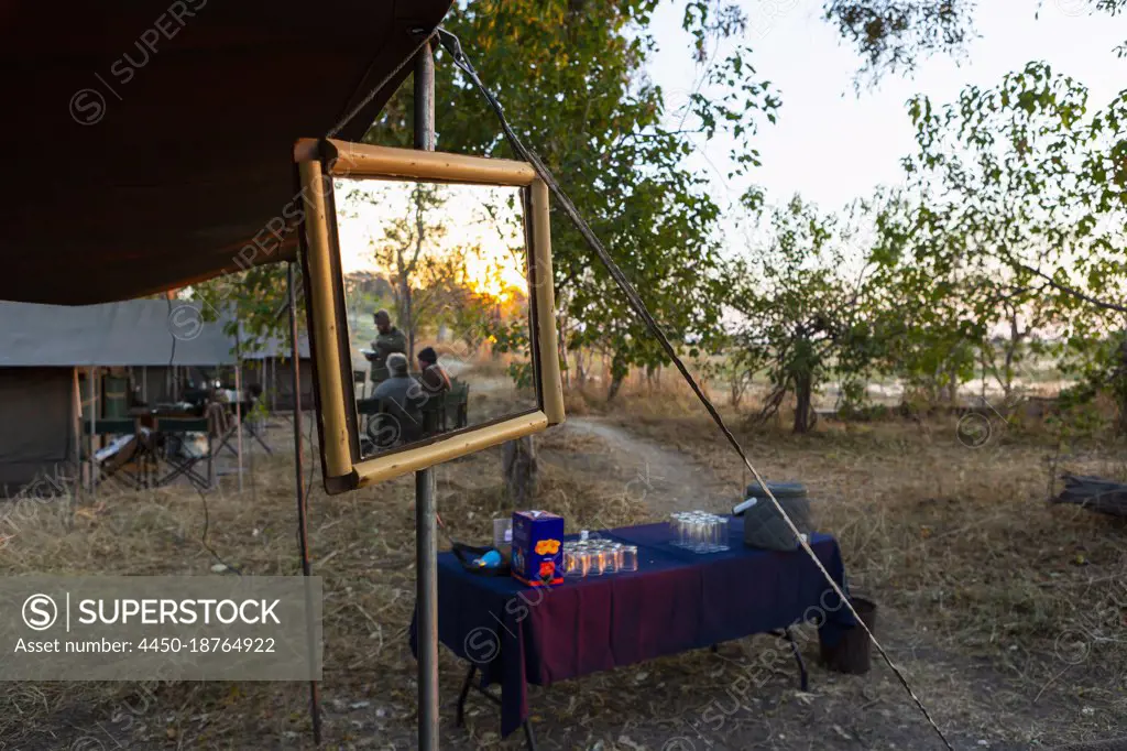 A safari camp, tents and drinks table, a large mirror on a tent pole, three people at sunset 