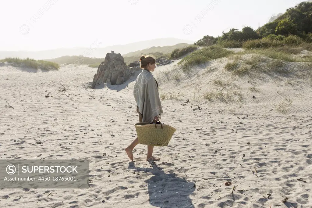 Teenage girl walking along the sand carrying a basket.