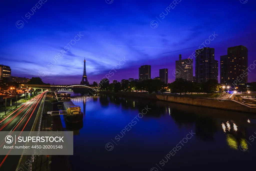 A view along the water of the River Seine at night, tall buildings on the river bank, the Eiffel tower in the distance. 