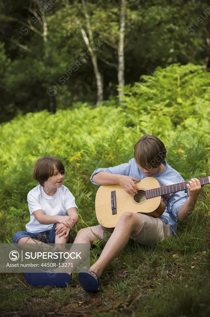 Two boys camping in New Forest. Sitting on the grass, one  playing a guitar.  Hampshire, England. 08/06/2013