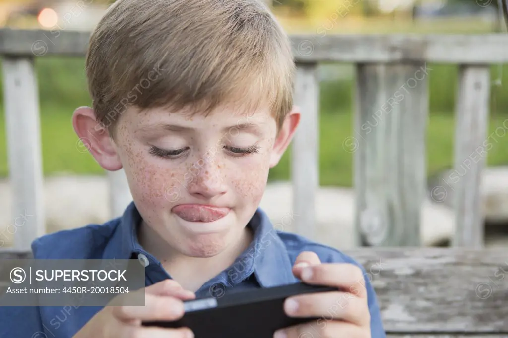 A young boy using a hand held electronic tablet or game and concentrating, sticking his tongue out.