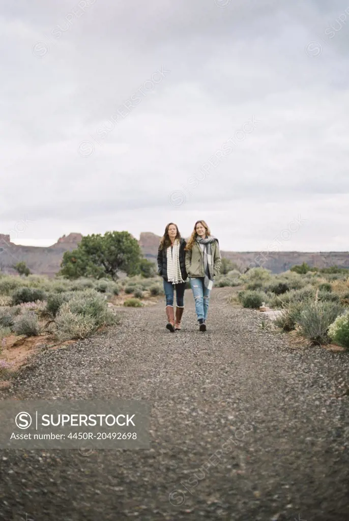 Two women walking side by side down a dirt road in a desert.