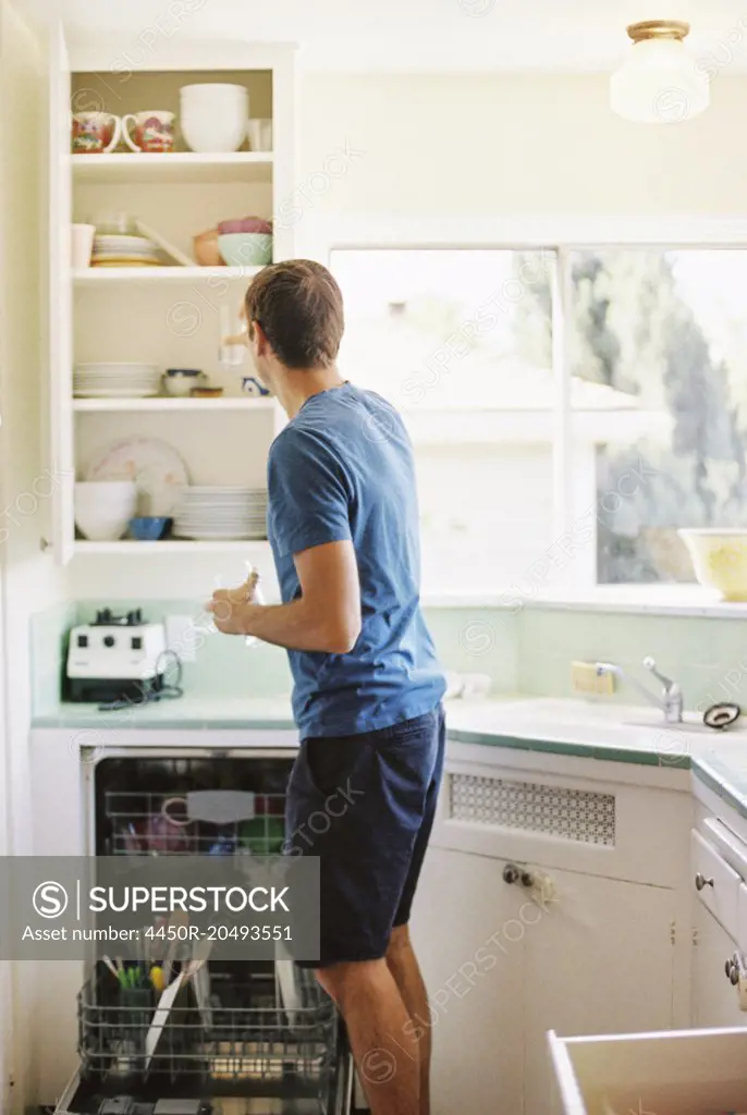 Rear view of a man standing in front of a cupboard with crockery in a kitchen.