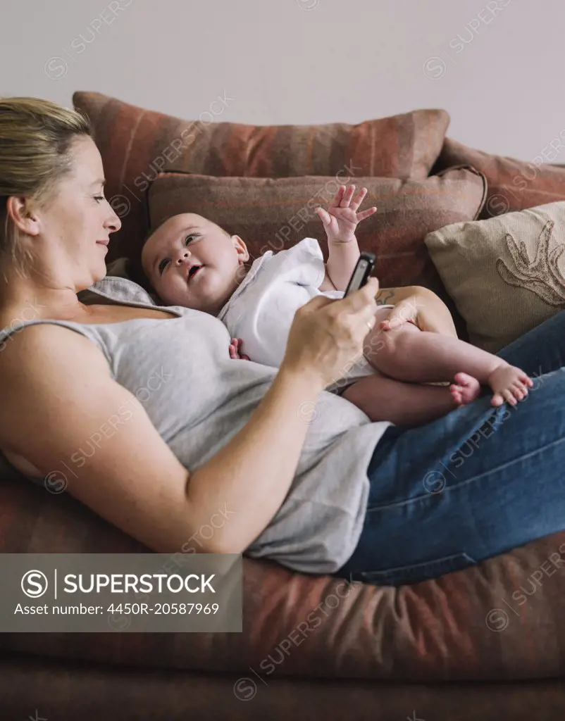 A woman lying on a sofa holding a baby girl, and holding a smart phone in one hand.