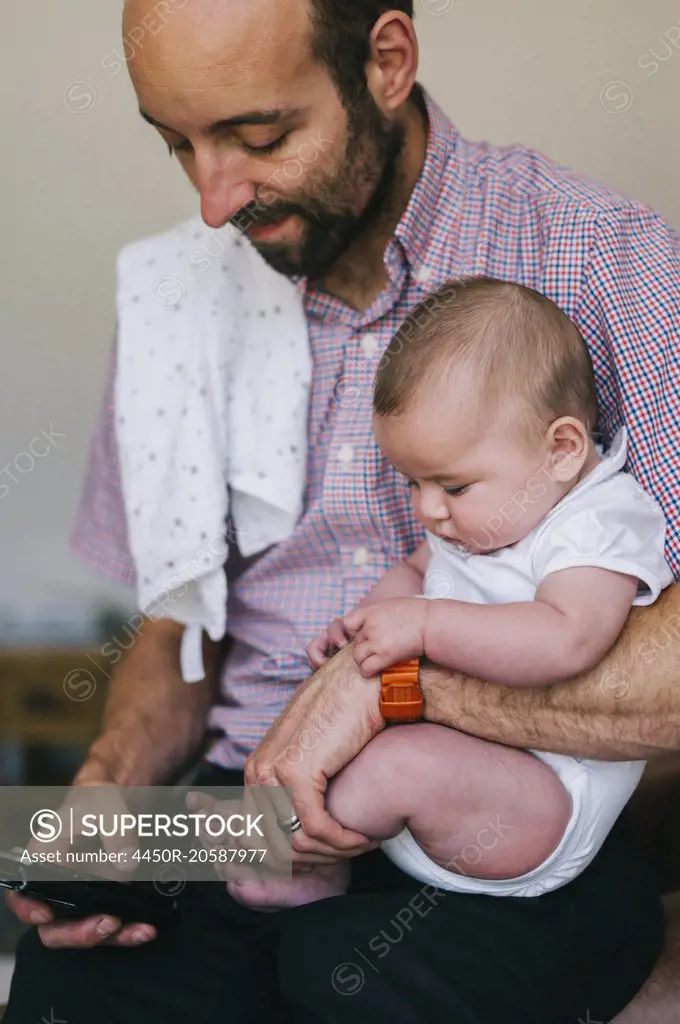 A man holding his small child in the crook of his arm, texting on his smart phone with the other.