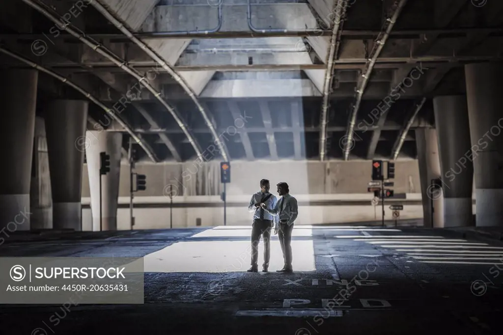 Two men in an urban underpass, with concrete and glass structures surrounding them, taking a selfie.