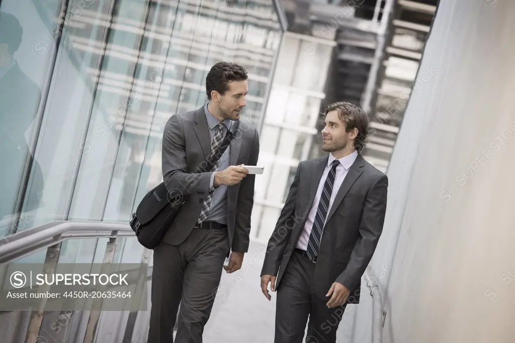 Two men in business suits outside a large building, one holding a smart phone.