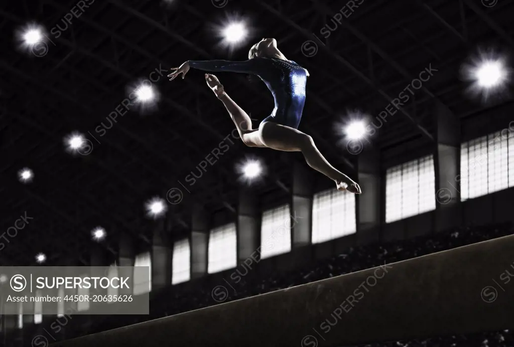 A female gymnast, a young woman performing a floor routine, in mid air with legs and arms outstretched and back bent.