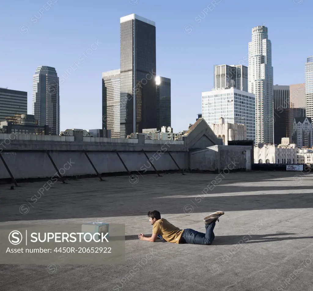 A man lying on his stomach on a city rooftop watching a small blue portable television.