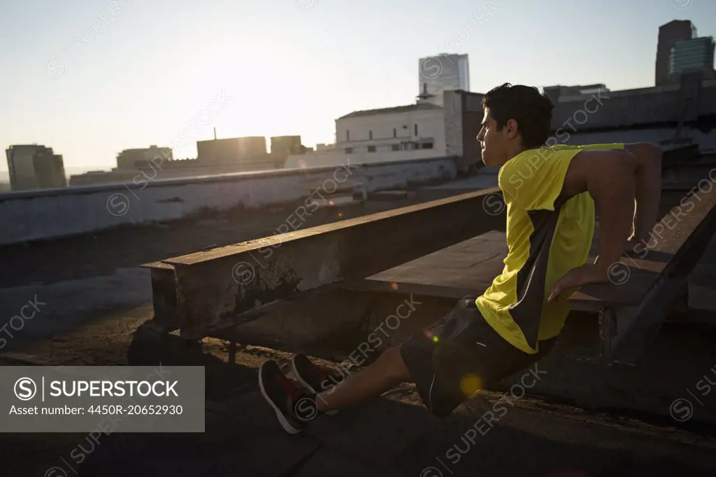 A man in exercise clothes on a rooftop overlooking the city, doing bench shoulder push ups.