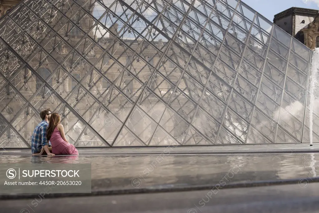 A couple in the courtyard of the Louvre museum, by the large glass pyramid. Fountains and water.