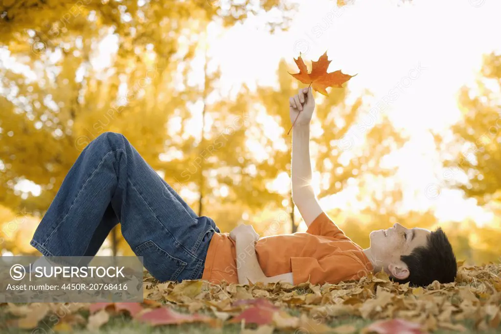 A boy lying on his back holding a maple leaf up to the autumn sun.