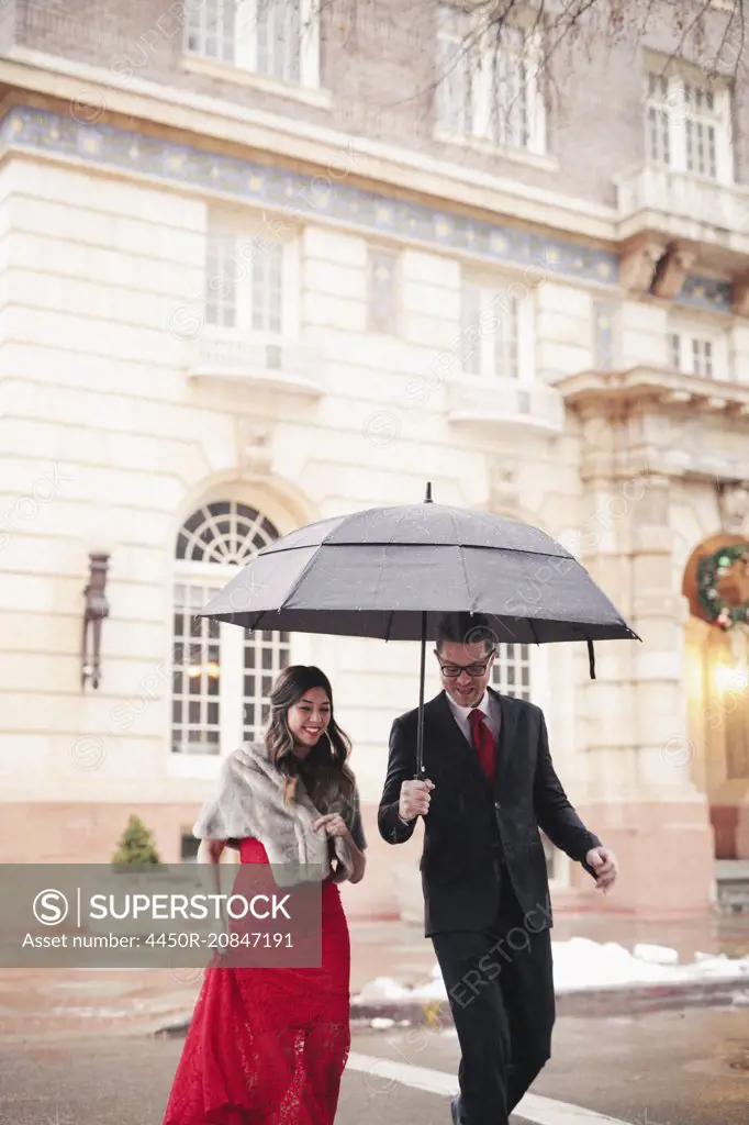 A woman in a long red evening dress with fishtail skirt and a fur stole, and a man in a suit, walking through snow in the city.