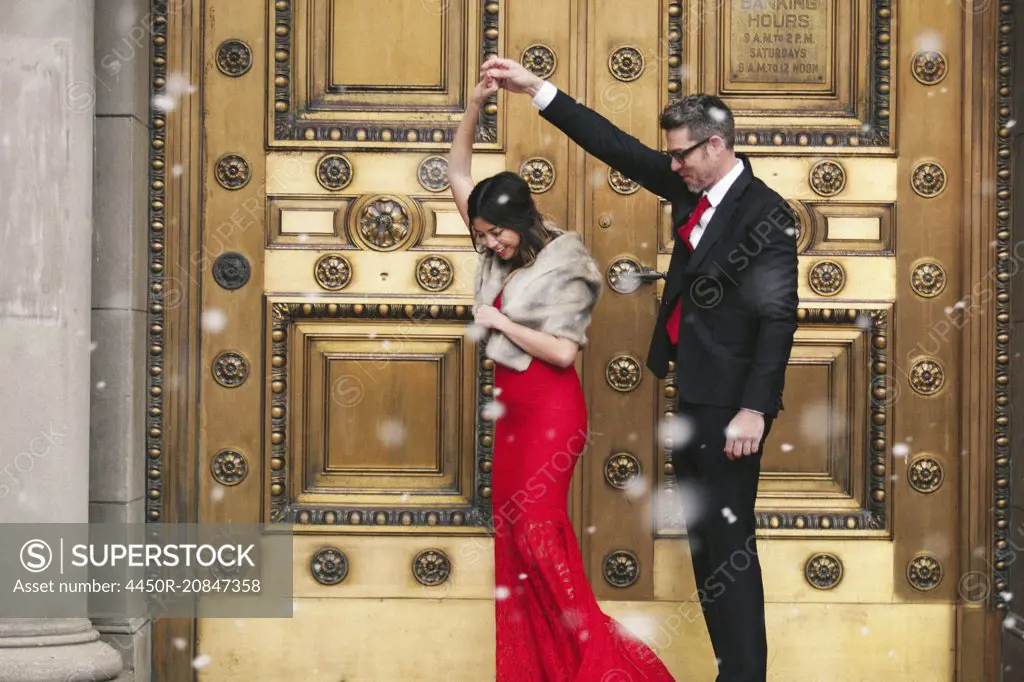 A woman in a long red evening dress with fishtail skirt and a fur stole, and a man in a suit dancing on the steps of a building.