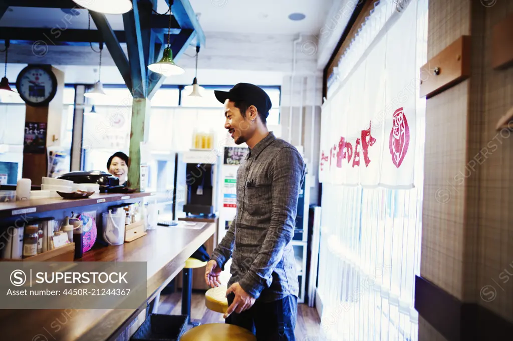 A ramen noodle cafe in a city.  Customer standing up at the counter ordering food.