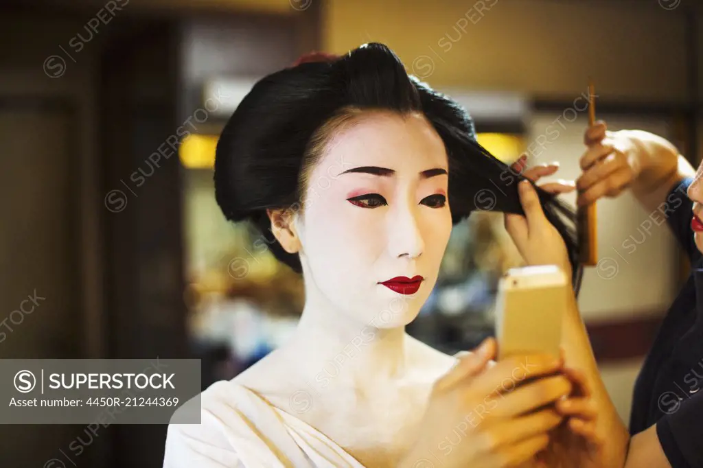 A modern geisha or maiko woman being prepared in traditional fashion, with white face makeup.