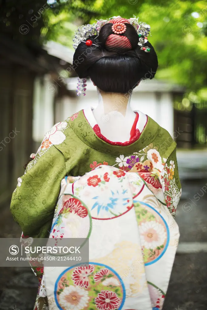 A woman dressed in the traditional geisha style, wearing a kimono and obi, with an elaborate hairstyle and floral hair clips, with white face makeup with bright red lips and dark eyes on a street.