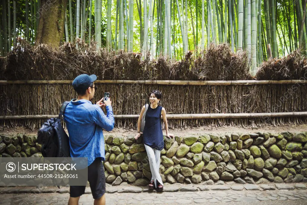 A man taking a photograph of a woman by a fence around woodland.