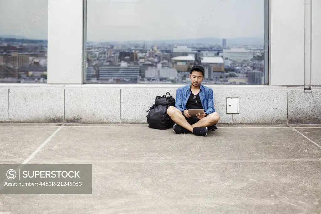 A man sitting on the floor using his smart phone, in front of a viewing point window on to a city.