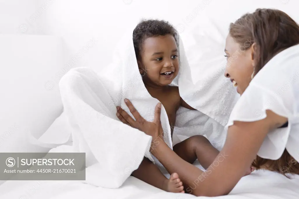 Women wrapping young boy sitting on a bed in white towel, looking at each other, smiling.