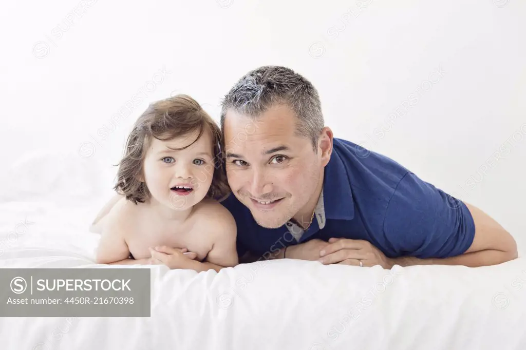 Man and young girl with brown hair lying side by side on a bed, smiling at camera.