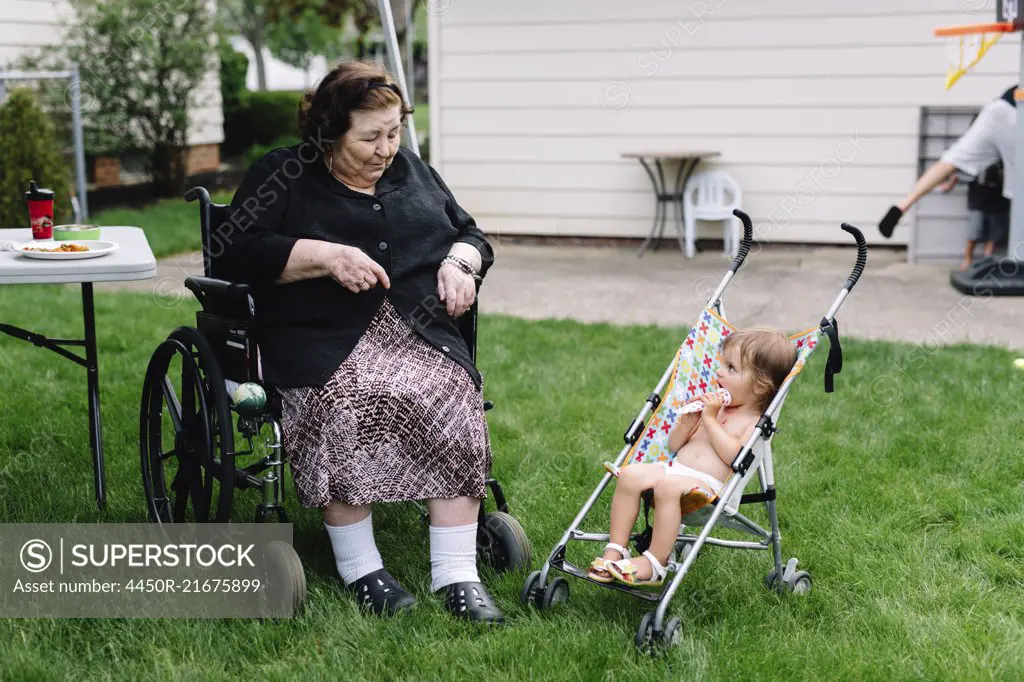 Smiling senior women sitting in a wheelchair in a garden beside bare chested young girl sitting in stroller.