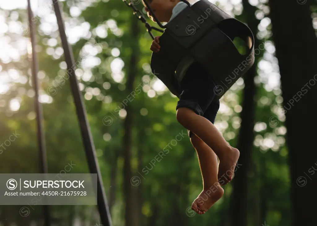 Low section view of barefoot boy sitting on a swing in a forest.